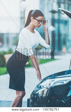 Examining her car. Side view of beautiful young businesswoman adjusting eyewear and looking inside of the vehicle hood while standing near her car