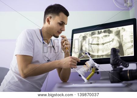 Intent dentist in a white uniform sits on the chair at the table. He looks at an articulator with teeth mould which he holds in the right hand. He holds pen in the left hand. On the table there is a monitor which shows the results of the orthopantomogram 