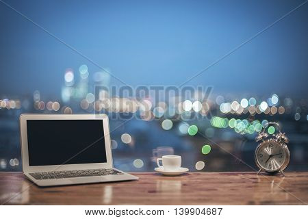Front view of creative designer desktop with blank laptop silver alarm clock and coffee cup on blurry night city background. Mock up