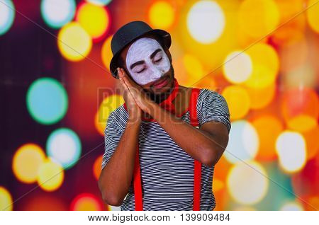 Headshot pantomime man with facial paint posing for camera using hands interacting sleeping, blurry lights background.