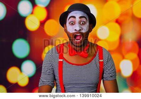 Pantomime man wearing facial paint posing for camera interacting making funny expressions, blurry lights background.