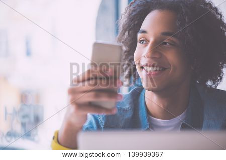 Modern one. Close up of smiling and cheerful young man holding his mobile phone and looking somewhere thinking about his plans
