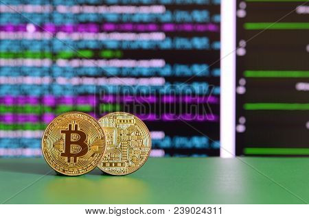 Two Gold Bitcoins Lie On The Green Surface On The Background Of The Display, Which Shows The Process
