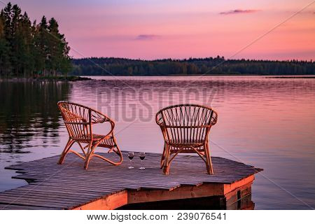 Two Wooden Chairs On A Wood Pier Overlooking A Lake At Sunset