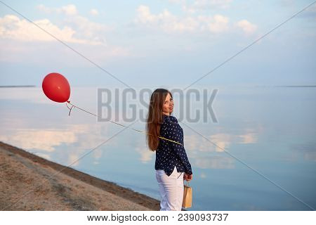 Portrait Of Young Woman With Red Air Balloon And Present Bag Near The Calm Sea Or Lake. Clouds Are R
