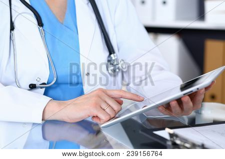 Woman Doctor Using Tablet Computer While Sitting At The Glass Table In Hospital Office, Closeup. Hea