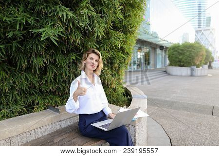 Female Manager Working With Laptop And Papers In Open Air Near Green Plant, Showing Thumbs Up. Conce