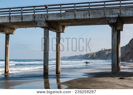 A Lone Male Surfer Carries A Surfboard Out To Sea Near A Concrete Pier In La Jolla, California