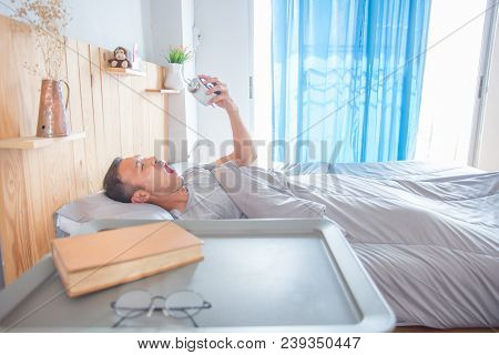 Man With Yawning Face Lies On Pillow, Holds Alarm Clock.