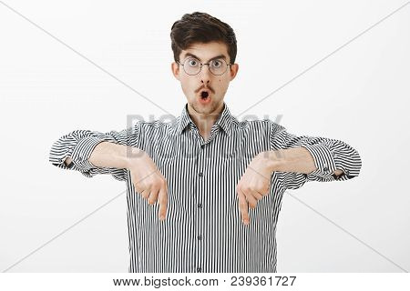 Studio Shot Of Shocked Amazed Creative Young Guy With Moustache In Glasses, Pointing Down With Index