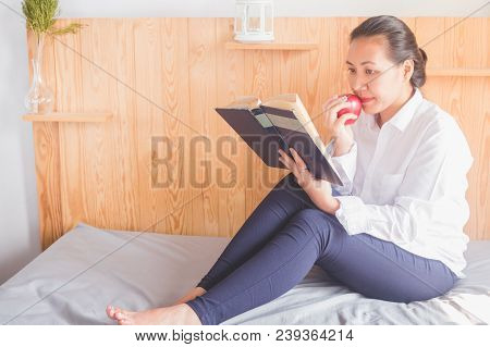 Asian Young Woman Reading Book And Eating Red Apple On Bed