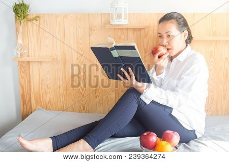 Asian Young Woman Reading Book And Eating Red Apple On Bed