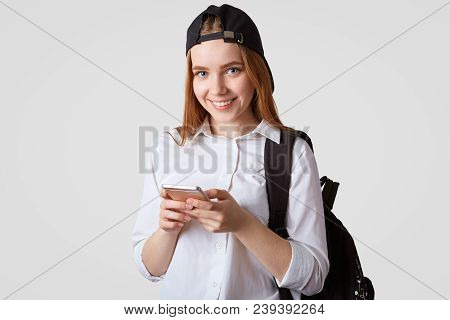 Isolated Shot Of Teenage Girl Comes Back From School, Carries Rucksack, Holds Modern Smart Phone, Su
