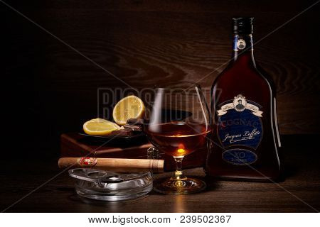 Still life with bottle of cognac Jacques Laffitte XO Superior, cigar Vasco da Gama on ashtray, glass with cognac and plate with pieces of lemon and chocolate stands on a table. Dark wooden background.