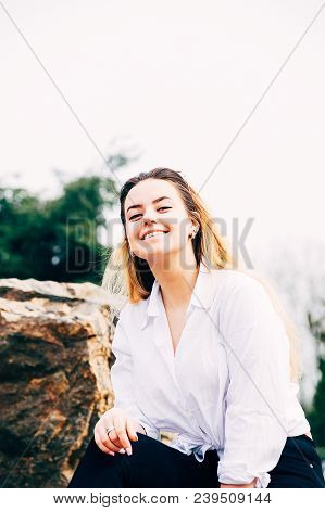 A Portrait Of A Pretty Young Long Haired Girl In White Shirt, Smiling, Sitting On A Big Stone In A P