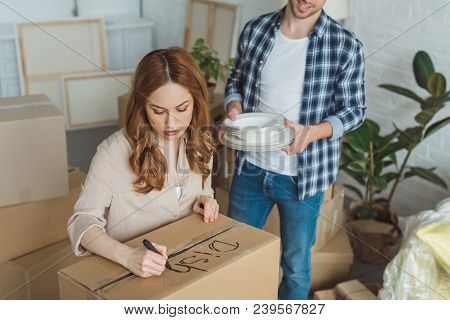 Partial View Of Woman Signing Cardboard Box With Husband With Dishes Near By, Moving Home Concept