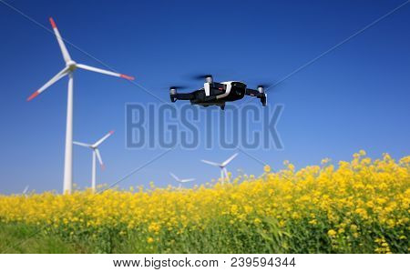 Inspection Drone With Eolian Turbines Behind. Rapeseed Field In Bloom. Renewable Energy.