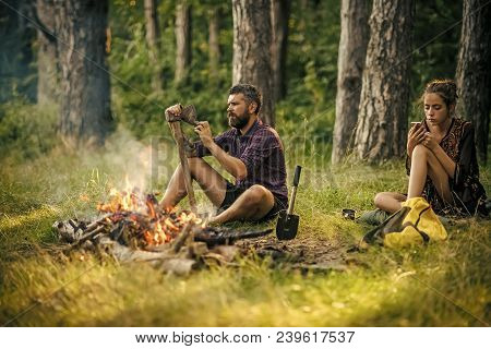 Couple Of Hikers Relax At Bonfire In Forest. Man Hipster Sharpen Axe. Girl Or Woman Use Mobile Phone