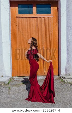 Beautiful Young Woman In A Long Red Evening Gown With A Train Standing Next To The Old Door