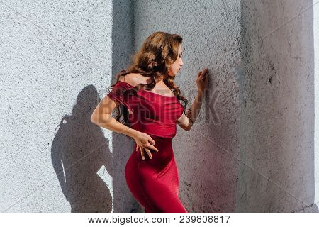 Beautiful Young Woman In A Long Red Evening Gown With A Train Standing Next To The Old Wall