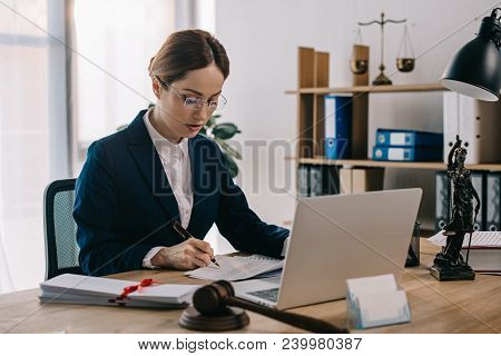 Side View Of Female Lawyer Doing Paperwork At Workplace With Laptop In Office