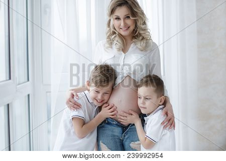 Two Boys, Brothers, Stand Next To A Beautiful Pregnant Mother,holding Hands On The Belly Of A Pregna