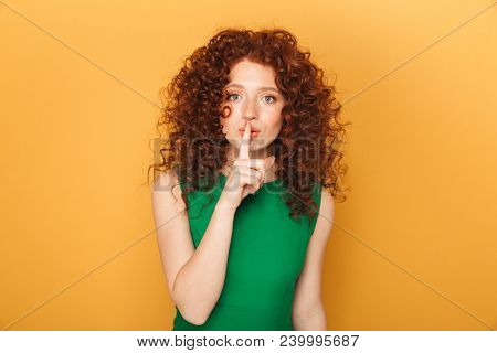 Portrait of a smiling curly redhead woman showing silence gesture isolated over yellow background