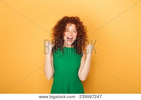 Portrait of a smiling curly redhead woman celebtrating success isolated over yellow background