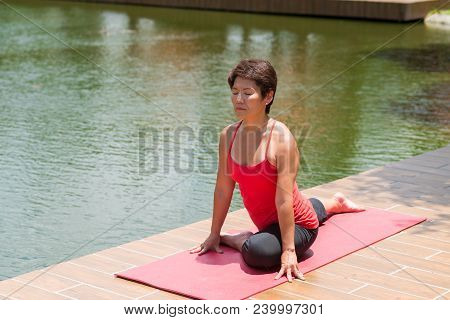 Japanese Mature Woman Sitting In Pidgeon Position By Pond In Park