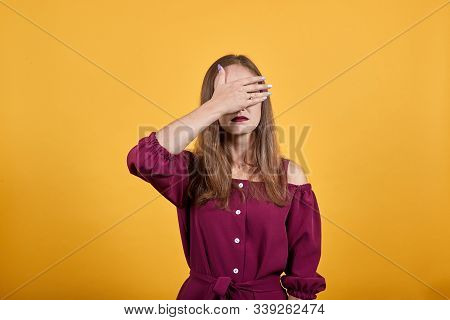 Girl In Burgundy Bluse Hides Face With Hand. Behind Her There Is Orange Background. Teen Looks Upset