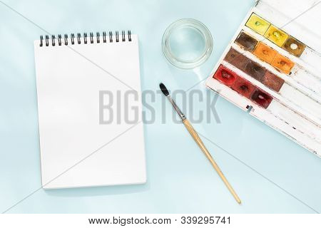 White Clean Note Pad On Black Spring, Squirrel Brush, Jar Of Water And Watercolor Paints On Blue Pap