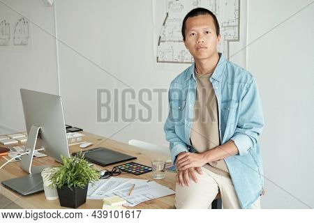 Young erious Chinese businessman sitting on table with papers, color palette, samples, graphic tablet and computer monitor