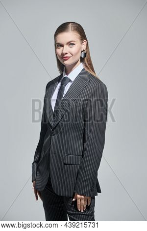 Fashion shot of a pretty young woman in a strict striped pantsuit posing smiling on a grey studio background. Impeccable look. 