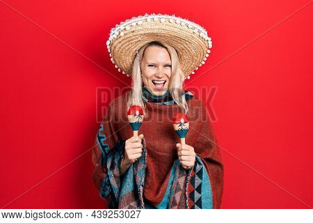 Beautiful caucasian blonde woman wearing festive mexican poncho and maracas smiling and laughing hard out loud because funny crazy joke. 