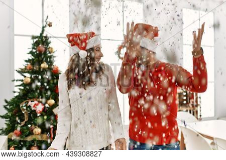 Young hispanic couple wearing christmas hat throwing conffeti celebrating new year at home.