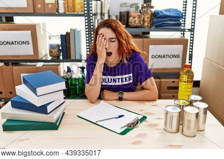 Young redhead woman wearing volunteer t shirt at donations stand yawning tired covering half face, eye and mouth with hand. face hurts in pain. 