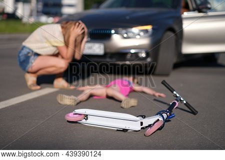Little Girl Lies Motionless On Asphalt With Scooter After Collision With Car.
