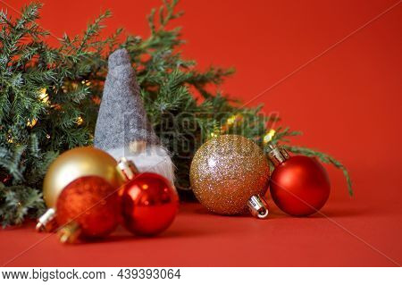 A Gnome In A Gray Hat Stands Next To A Spruce Branch And Many Red And Gold Christmas Balls