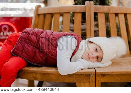 Tired Boy In White Bobble Hat Is Lies With His Mittened Hands Under His Head On A Wooden Bench In Sh