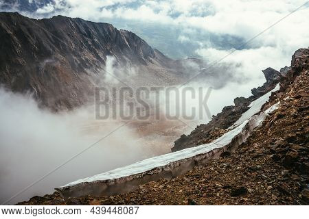 View From Precipice Edge With Sharp Rocks And Snow Cornice To Mountain Valley In Low Clouds. Wonderf