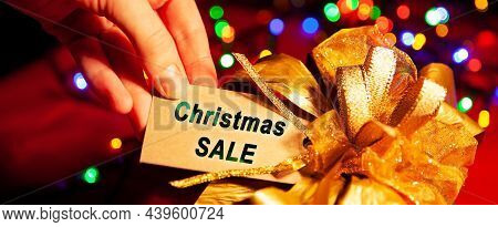 A Woman's Hand Holds A Label With Text - Christmas Sale, On A Gift Box With A Gold Bow, Close-up. On