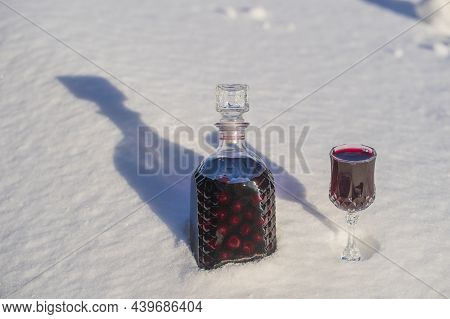 Homemade Tincture Of Red Cherry In A Glass Bottle And A Wine Crystal Glass On A Snow And White Backg