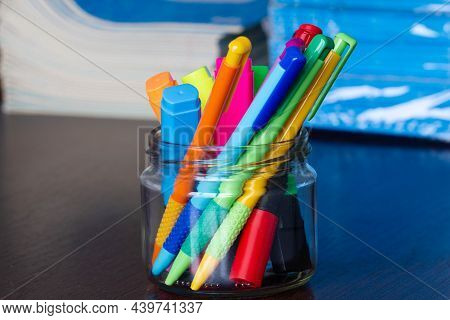 Multi-colored Pens And Markers In A Stand On The Background Of Stack Of Notebooks.  Pack Of Blue Sch
