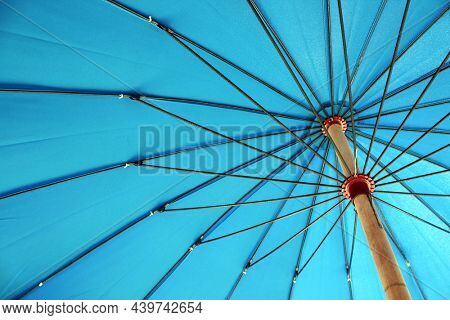 Under a blue parasol showing the details of the metal spokes. The umbrella is providing sun block yet projecting a vivid aqua blue background color.