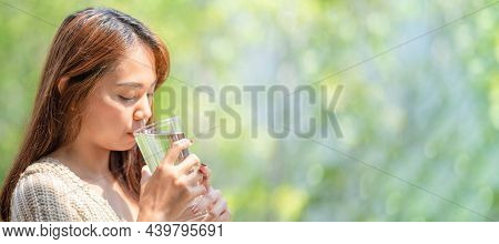 Asian Woman Drinks Water From Tall Glass Of Water At Outdoor Tree Bokeh Green For Banner Background.