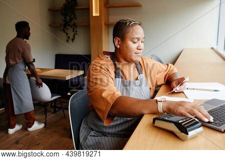 Young African American Woman Wearing Apron Sitting At Table In Front Of Laptop In Modern Cafe Taking