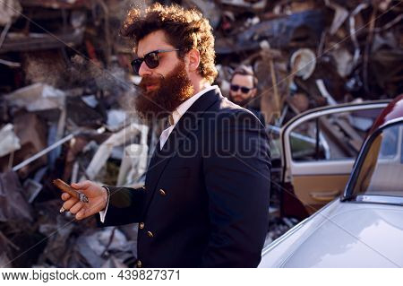 Stylish Reddish Hair, Bearded Man Smokes A Cigar On A Old Iron Dump, Wearings In Suit And Black Sung
