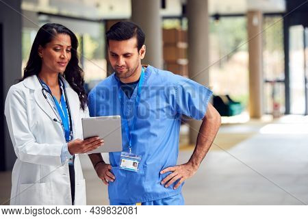 Two Medical Staff In White Coats And Scrubs With Digital Tablet Having Informal Meeting In Hospital