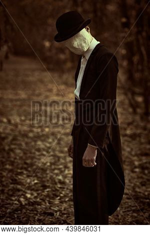 A strange and creepy gentleman in a mask of plaster bandage on his face, in a bowler hat and a black tailcoat, stands in an old autumn park. Halloween. Vintage style. Horror, thriller.