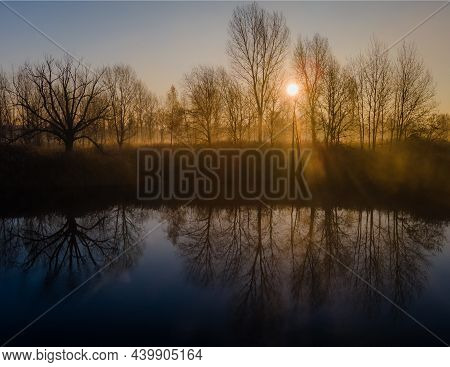 The Rays Of The Morning Sun Shining Through The Autumn Trees And Reflected In The Water Of The Misty